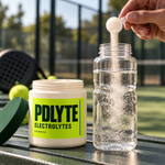PDLYTE electrolytes preparation: a hand tipping a measuring scoop into an open water bottle beside the open powder pot